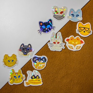 May include: A collection of holographic cat-themed stickers in various colors and designs. The stickers feature cat faces with different expressions, some wearing accessories like glasses or scarves. The stickers are arranged on a white and brown background.