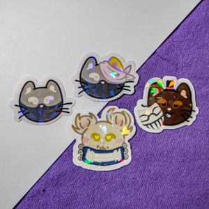 以下が含まれることがあります： 動物の漫画キャラクターが描かれた、虹色のホログラフィックステッカー4枚。 ステッカーは白と紫の表面に貼られています。 キャラクターには、目隠しをした猫、シルクハットをかぶった猫、包帯をした猫、青い首輪をした犬が含まれています。