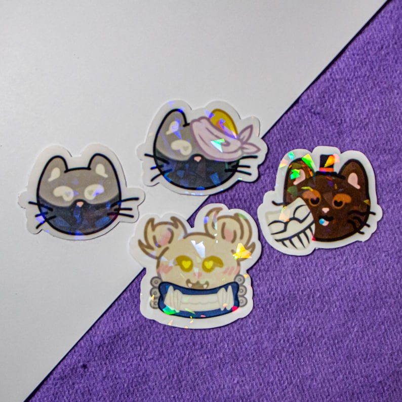 以下が含まれることがあります： 動物の漫画キャラクターが描かれた、虹色のホログラフィックステッカー4枚。 ステッカーは白と紫の表面に貼られています。 キャラクターには、目隠しをした猫、シルクハットをかぶった猫、包帯をした猫、青い首輪をした犬が含まれています。