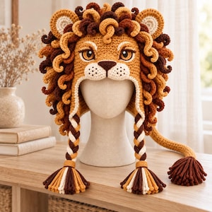 May include: A crocheted lion hat featuring a golden-yellow face, a thick mane of curly brown and tan yarn, and two long braided earflaps ending in multi-colored tassels. The hat includes a long tail extension with a dark brown pom-pom.