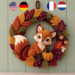 Op de afbeelding: Een gehaakte herfstkrans met een centrale oranje en witte vossenfiguur, een kleine oranje pompoen, eikels, een paddenstoel en diverse herfstkleurige bladeren in rood, oranje, geel en groen, gerangschikt op een donkerbruine basis.
