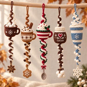 Könnte beinhalten: Fünf gehäkelte Hängedekorationen mit Tassen und Gläsern, verziert mit Schlagsahne, Zimtstangen und Beeren. Die Designs zeigen Schneeflocken, ein Herz und das Wort COCOA, ergänzt durch baumelnde Anhänger wie Sterne, Löffel und Schneeflocken.