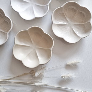 May include: Five small, white ceramic bowls with a flower-like shape. The bowls have a textured surface and are arranged on a white background.