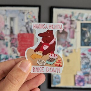 Puede incluir: Una pegatina con un oso de dibujos animados horneando. La pegatina dice "HAVING A MENTAL BAKE DOWN" con imágenes de galletas y cupcakes. El oso lleva un delantal rojo y un cuello blanco con volantes.