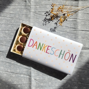 Könnte beinhalten: Eine weiße Geschenkbox mit dem Wort "DANKESCHÖN" in bunten Buchstaben. Die Box enthält mehrere runde Pralinen. Die Box ist mit kleinen gelben Blumen verziert.