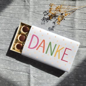 Könnte beinhalten: Eine weiße Geschenkbox mit dem Wort "DANKE" in bunten Buchstaben. Die Box enthält mehrere runde Pralinen mit weißer Füllung. Die Box liegt auf einem grauen Stoff mit Blumenmuster, mit einem Zweig getrockneter Blumen.