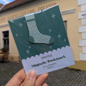 Könnte beinhalten: Ein handgefertigter magnetischer Lesezeichen mit einem grünen Sockenmuster und weißen Sternakzenten. Auf dem Lesezeichen steht "HANDMADE Magnetic Bookmark". Das Lesezeichen wird in einer Hand gehalten.