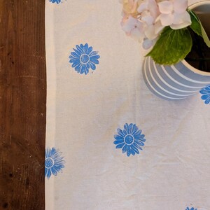 Può includere: Tessuto bianco con un motivo di margherite blu. Le margherite sono uniformemente distribuite sul tessuto. Un vaso con fiori rosa e foglie verdi è nell'angolo in alto a destra. Il tessuto è su una superficie di legno.