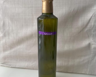 May include: A tall, green glass bottle with a gold-coloured cap. A purple label with the word "Spikenard" is affixed to the bottle. The bottle is filled with a dark liquid.