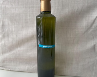May include: A tall, green glass bottle with a gold-colored cap and a blue label that reads "Frankincense." The bottle is filled with a dark liquid. The bottle is approximately 10 inches tall.