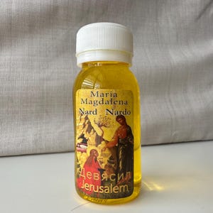 May include: A small glass bottle filled with yellow liquid. The label features a painting of Mary Magdalene and the text "Maria Magdalena Nard Nardo Девясил Jerusalem".