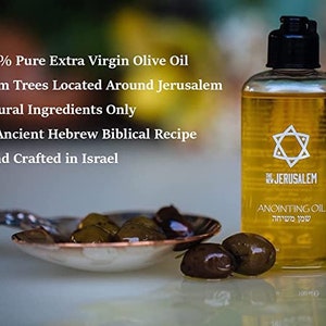 May include: A clear glass bottle of anointing oil with a gold star of David on the label. The bottle is labeled "The New Jerusalem Anointing Oil" in English and Hebrew. The bottle is sitting on a table with olives in the foreground.