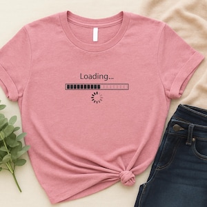 Könnte beinhalten: Ein rosa T-Shirt mit dem Text "Loading..." über einer Fortschrittsbalken-Grafik. Das Shirt wird mit dunkelblauen Jeans kombiniert. Ein Zweig mit grünen Blättern ist sichtbar.