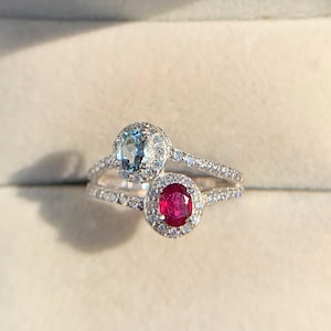 May include: Two silver rings with gemstone settings. One ring features a light blue gemstone, and the other has a red gemstone. Both rings are set with smaller, clear gemstones. The rings are displayed in a red jewelry box.