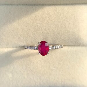 Può includere: Anello in argento con una grande pietra centrale di rubino ovale. La fascia è impreziosita da piccoli diamanti scintillanti. L'anello è presentato in un cofanetto nero con interno color crema.