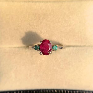 May include: A silver ring featuring a central oval-cut red ruby flanked by two smaller round-cut green emeralds. The stones are set in a prong setting on a polished silver band, creating a three-stone design with a vibrant color contrast.