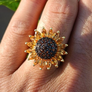 Puede incluir: Un anillo de oro con un diseño de girasol. El girasol está hecho de piedras preciosas amarillas y marrones.