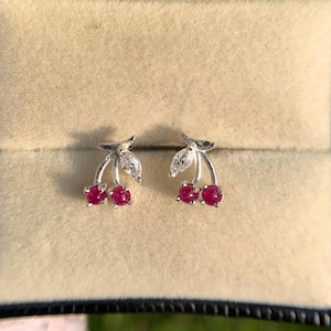 May include: A pair of silver stud earrings shaped like cherries, featuring two round, deep pink stones as the fruit and a marquise-cut clear stone as a leaf on each earring. The earrings are displayed side by side in a beige velvet jewellery box.