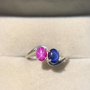 Op de afbeelding: Een zilveren ring met twee ovale edelstenen, één roze en één blauwe. De edelstenen zijn naast elkaar geplaatst in een wervelend ontwerp. De ring wordt gepresenteerd in een rode en beige sieradendoos.