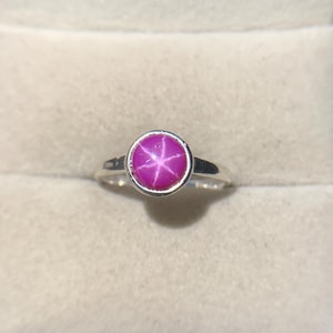 May include: A silver ring with a round, bright pink gemstone displaying a six-pointed star. The ring has a bezel setting and a plain, polished band. It is presented in a red jewellery box.