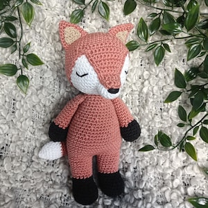 May include: A crocheted fox plush toy with closed eyes, a white muzzle, and black feet. The toy is lying on a white fluffy surface.