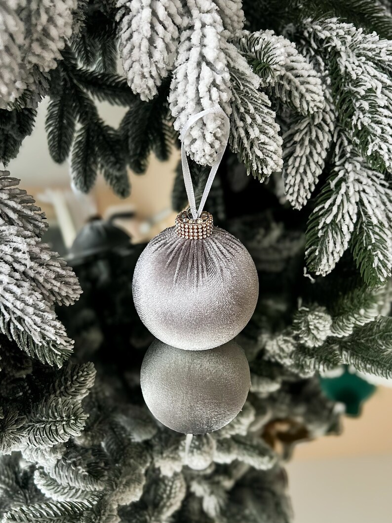 Puede incluir: Un adorno navide&ntilde;o de terciopelo plateado con ribete de cuentas cuelga de un &aacute;rbol de Navidad artificial nevado.