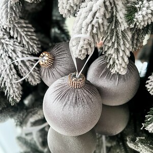 Puede incluir: Tres adornos navide&ntilde;os de terciopelo gris con adornos dorados y de cristal colgados de una rama de &aacute;rbol de Navidad artificial cubierta de nieve.