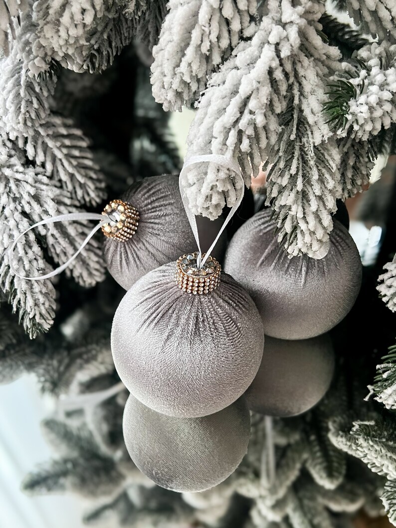 Puede incluir: Tres adornos navide&ntilde;os de terciopelo gris con adornos dorados y de cristal colgados de una rama de &aacute;rbol de Navidad artificial cubierta de nieve.