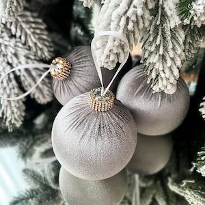 Puede incluir: Tres adornos navide&ntilde;os de terciopelo gris con detalles de diamantes de imitaci&oacute;n dorados y transparentes colgados de un &aacute;rbol de Navidad artificial nevado.