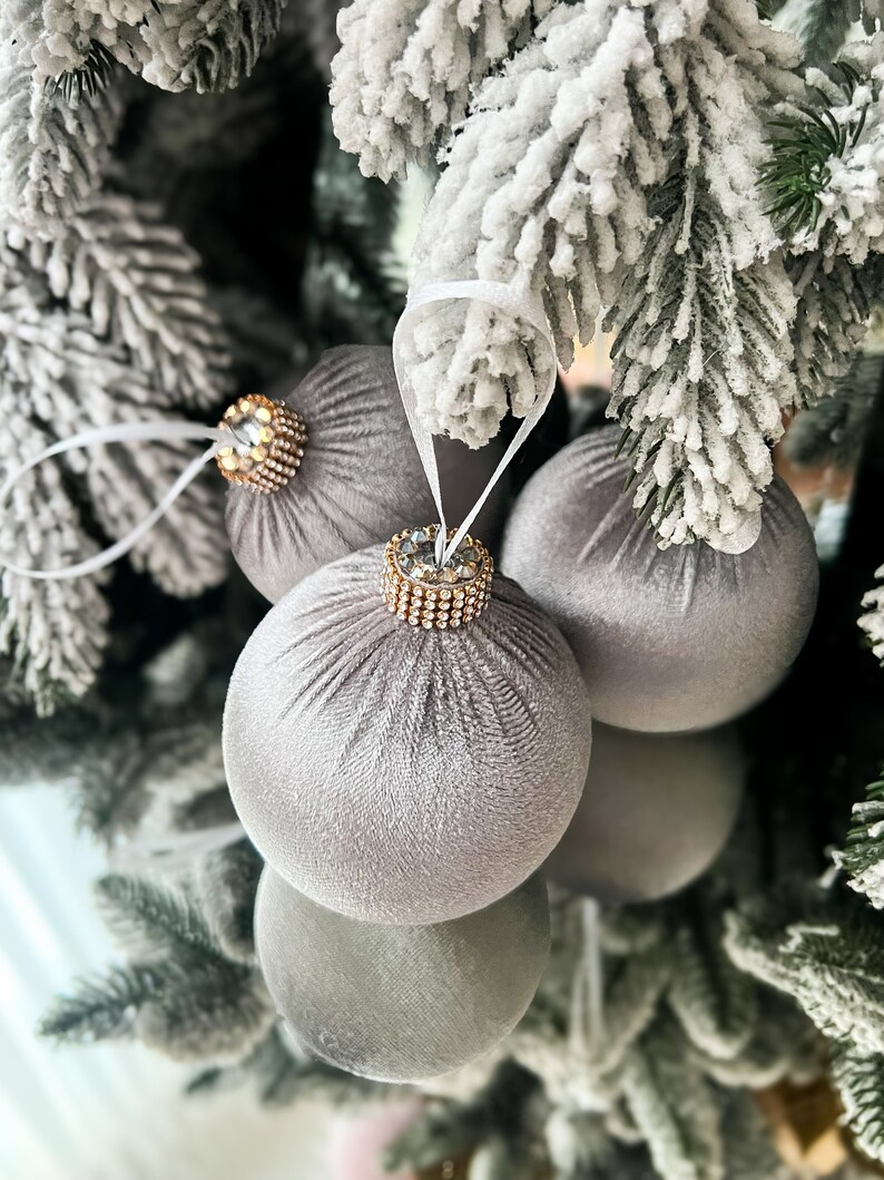 Puede incluir: Tres adornos navide&ntilde;os de terciopelo gris con detalles de diamantes de imitaci&oacute;n dorados y transparentes colgados de un &aacute;rbol de Navidad artificial nevado.