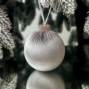 Puede incluir: Un adorno navide&ntilde;o de terciopelo plateado con ribete de diamantes de imitaci&oacute;n dorados. El adorno cuelga de una cinta blanca en una rama de pino cubierta de nieve.