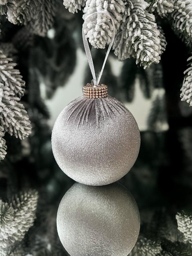 Puede incluir: Un adorno navide&ntilde;o de terciopelo plateado con ribete de diamantes de imitaci&oacute;n dorados. El adorno cuelga de una cinta blanca en una rama de pino cubierta de nieve.