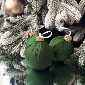May include: Three spherical ornaments wrapped in dark green fabric with vertical pleats. Each ornament features a gold-toned cap encrusted with small clear crystals and a white ribbon loop for hanging, reflecting on a smooth surface below.