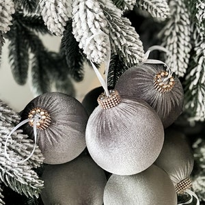 Puede incluir: Un conjunto de seis adornos navide&ntilde;os de terciopelo gris con ribete de cuentas. Los adornos est&aacute;n colgados de un &aacute;rbol de Navidad artificial nevado.