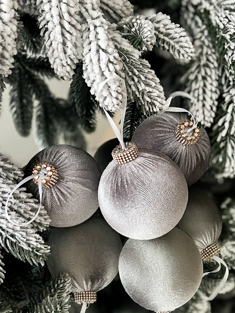 Puede incluir: Un conjunto de seis adornos navide&ntilde;os de terciopelo gris con ribete de cuentas. Los adornos est&aacute;n colgados de un &aacute;rbol de Navidad artificial nevado.