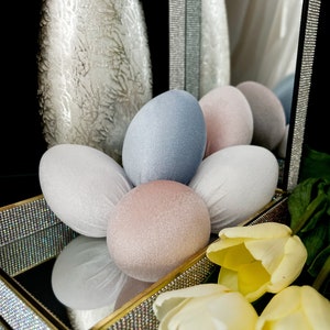 Pode incluir: Arranjo decorativo com ovos de veludo em tons pastel, azul, rosa e branco, sobre uma bandeja espelhada com borda dourada. Tulipas amarelas e um vaso prateado completam a cena.