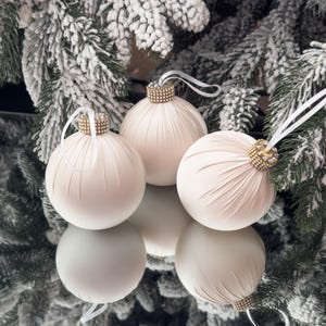 Op de afbeelding: Drie ivoorkleurige kerstballen met geplooide stoffen details en juwelen doppen. Elke ornament heeft een wit lint om op te hangen. De ornamenten worden gepresenteerd voor een bevroren dennenboom.