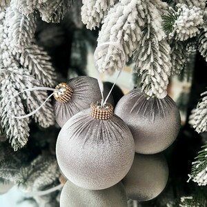Puede incluir: Tres adornos navide&ntilde;os de terciopelo gris con detalles dorados y de pedrer&iacute;a colgando de una rama de pino cubierta de nieve.