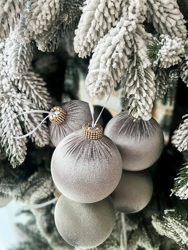 Puede incluir: Tres adornos navide&ntilde;os de terciopelo gris con detalles dorados y de pedrer&iacute;a colgando de una rama de pino cubierta de nieve.