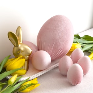 Könnte beinhalten: Osterdekoration mit goldener Hasenfigur, rosa Samteiern und gelben Tulpen. Der Hase steht auf einem weißen Tablett mit dekorativem Rand, neben einem großen Ei und mehreren kleineren. Tulpen im Vordergrund.