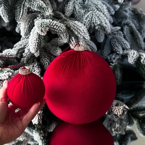 Puede incluir: Dos adornos navideños de terciopelo rojo con un borde brillante. El adorno más grande está en una rama de pino nevada, mientras que el adorno más pequeño se sostiene en una mano.