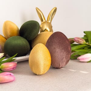 Könnte beinhalten: Eine Osterdekoration mit dekorativen Velveteiern in Gelb, Grün und Braun. Eine goldene Hasenfigur steht hinter den Eiern. Rosa Tulpen und grüne Blätter sind auf einer neutralfarbenen Tischdecke arrangiert.