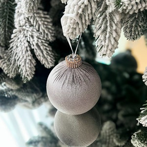 Puede incluir: Un adorno navide&ntilde;o plateado con un borde dorado brillante cuelga de una rama de &aacute;rbol de Navidad artificial cubierta de nieve.