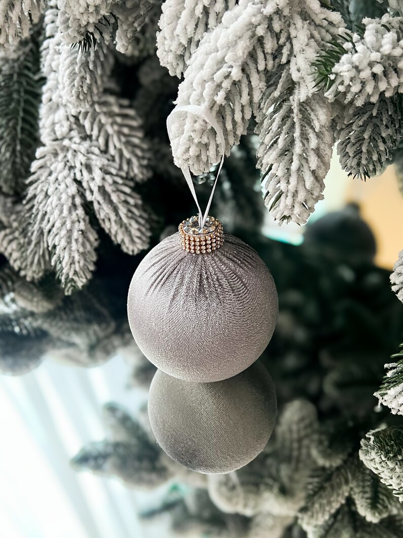 Puede incluir: Un adorno navide&ntilde;o plateado con un borde dorado brillante cuelga de una rama de &aacute;rbol de Navidad artificial cubierta de nieve.