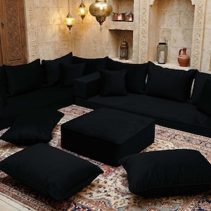 May include: A black modular sofa set with multiple cushions and a square ottoman. The furniture is arranged on a patterned rug, with decorative lamps and wall niches in the background. The overall style is a Middle Eastern or Moroccan theme.