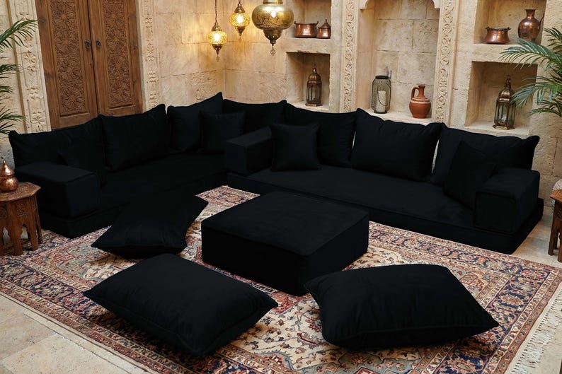 May include: A black modular sofa set with multiple cushions and a square ottoman. The furniture is arranged on a patterned rug, with decorative lamps and wall niches in the background. The overall style is a Middle Eastern or Moroccan theme.