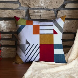 May include: Square decorative pillow with a geometric pattern in white, red, orange, yellow, gray, and blue. The pillow is placed on a wooden surface in front of a stone wall. A cream-colored blanket is draped nearby.