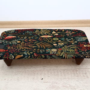May include: A rectangular footstool with a dark green floral pattern. The design includes birds, flowers, and foliage in shades of red, yellow, and green. The stool is supported by four dark wooden legs.