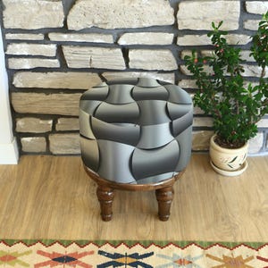 Peut inclure: Un petit pouf rond avec un motif abstrait gris et argenté sur le dessus rembourré. Le pouf a des pieds en bois tourné brun foncé. Une petite plante en pot est à proximité contre un mur de pierre.