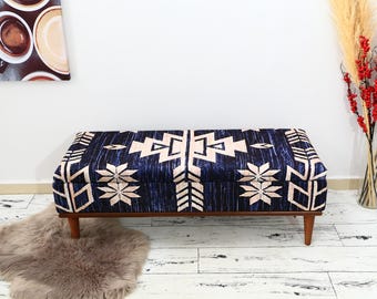 May include: A rectangular ottoman with a dark blue and cream geometric pattern. The ottoman has a wooden frame and four tapered legs. The design features star and arrow shapes. A fluffy, grey rug is in front of the ottoman.